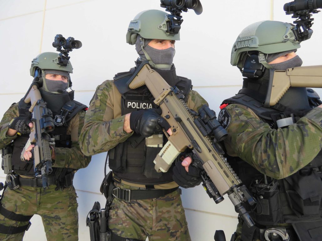 Tres agentes uniformados, con cascos y armas largas, posan frente a una pared clara.