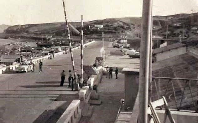Imagen en blanco y negro de un antiguo paso fronterizo, con barreras, vehículos y varias personas.