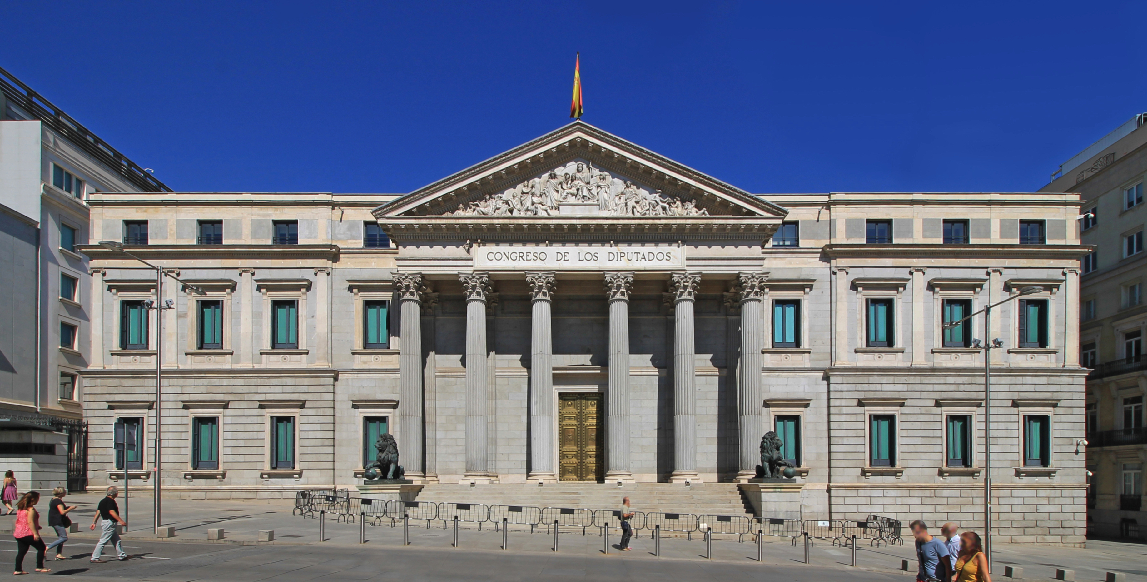 Fachada de un edificio institucional con bandera española en lo alto y varias personas en el exterior.